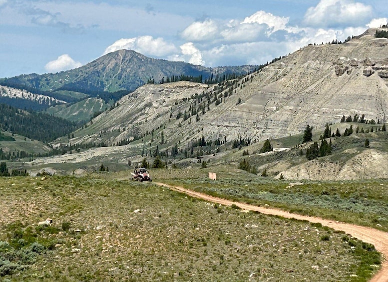 A jeep on a dirt road in a mountainous area with sparse vegetation and distant snow-capped peaks. 4