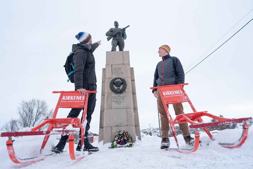 Two people standing on sleds with 'KIRKENES' written on them, facing a monument with a wreath and a statue. 4