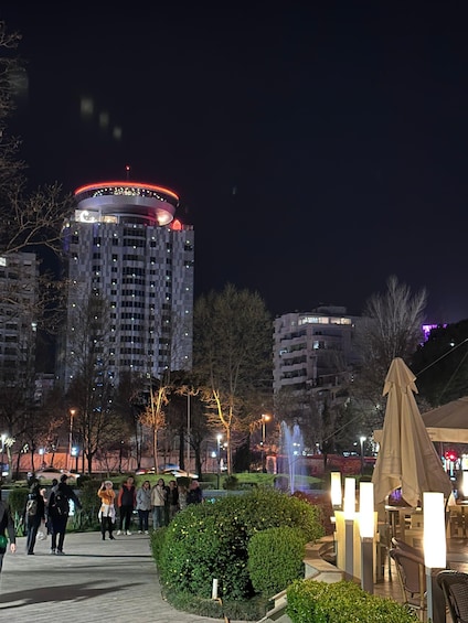 A nighttime urban scene with a brightly lit building, a fountain, and people walking. 4