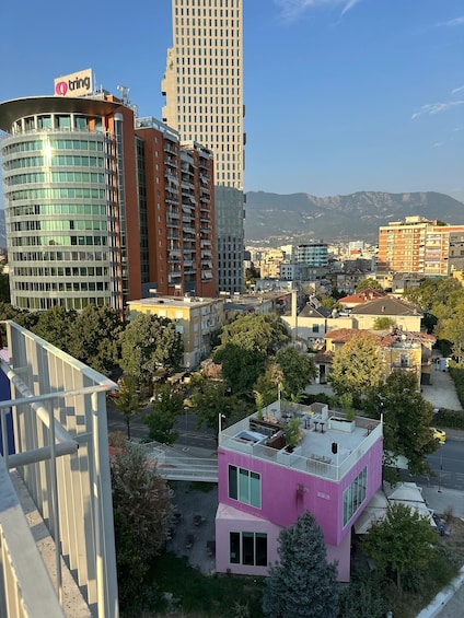 A cityscape with modern buildings, including a pink structure with outdoor seating, surrounded by trees and a mountain range in the background. 2