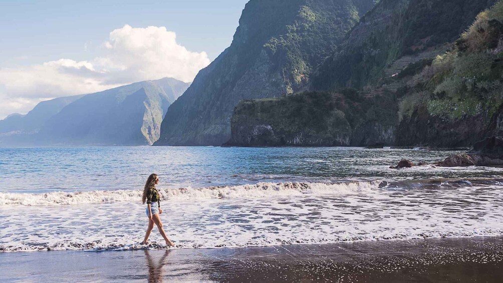 A woman walking along a beach with waves crashing onto the shore, surrounded by steep cliffs and mountains in the background. 5