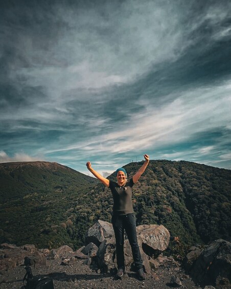 A person standing on a rocky outcrop with arms raised, overlooking a forested mountain under a cloudy sky. 4