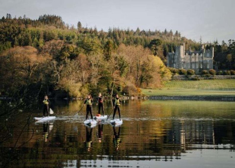 Four people paddleboarding on a calm lake with a forested hill and a castle in the background. 1