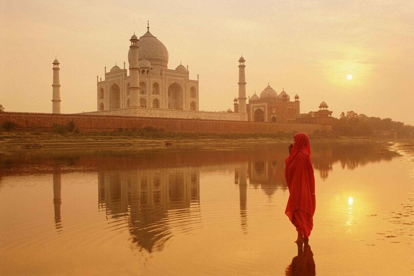 A person in red standing by a calm river reflecting a large white building with domes and minarets. 2