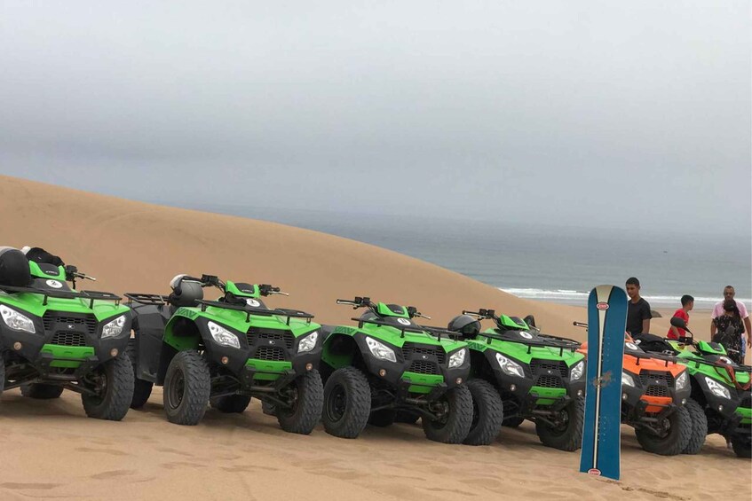 A row of green and orange quad bikes lined up on a sandy beach with a blue surfboard leaning against one of them. 1