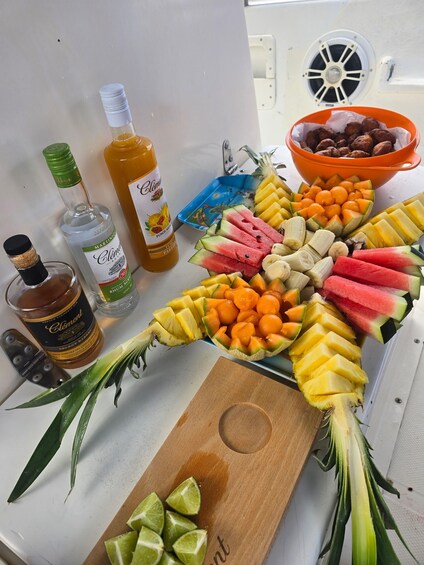 A table with a fruit arrangement, bottles of Clément and other beverages, and a wooden cutting board with lime slices. 2
