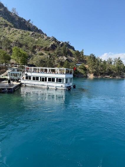 A white boat docked at a pier with people on board, surrounded by clear blue water and a mountainous landscape. 2
