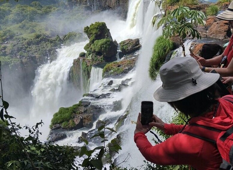 A waterfall with lush greenery and a person in a red jacket taking a photo. 3