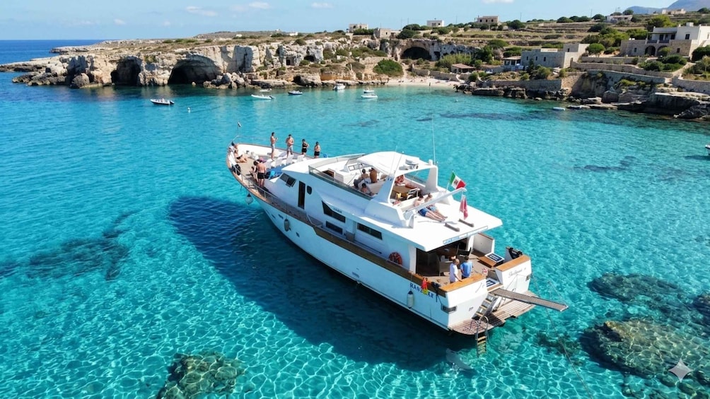 A white yacht docked in clear turquoise waters with rocky coastline and traditional buildings in the background. 1