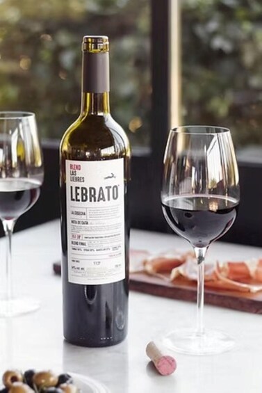 A bottle of LEBRATO wine, a glass of red wine, and a corkscrew on a table with a window view. 1