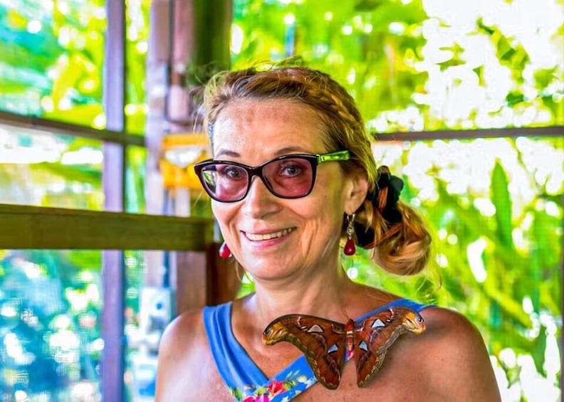 A person with a butterfly-shaped necklace and red earrings, standing in a green, leafy environment. 5