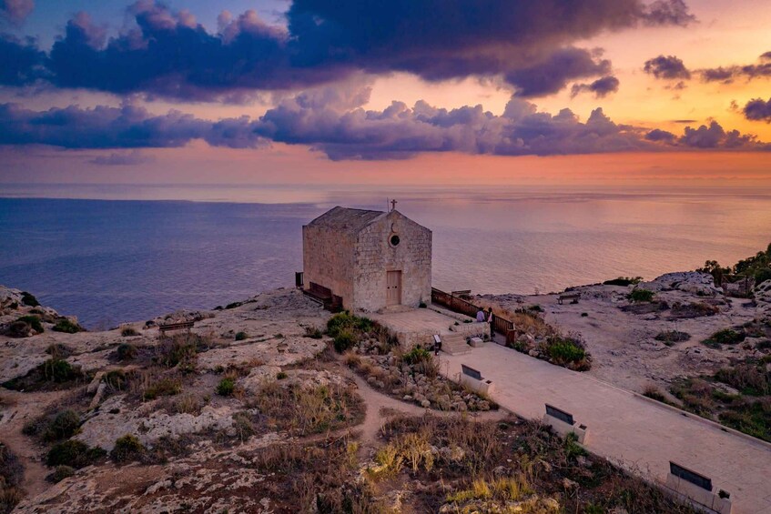 A coastal church with a stone facade, a wooden fence, and a clear view of the sea at sunset. 4
