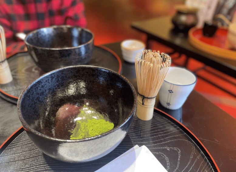 A traditional Japanese tea ceremony setting with a black bowl containing green powdered tea, a whisk, and a white cup. 4