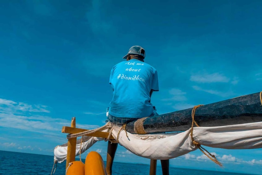 A person wearing a blue shirt with text on the back, standing on a boat with a rope and wooden structure. 2