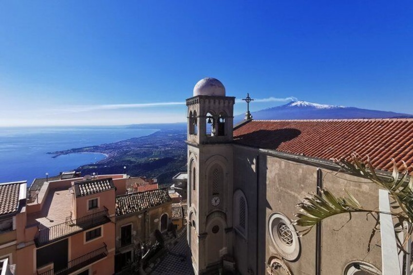 A coastal town with a prominent church tower, terracotta rooftops, and a snow-capped mountain in the background. 1