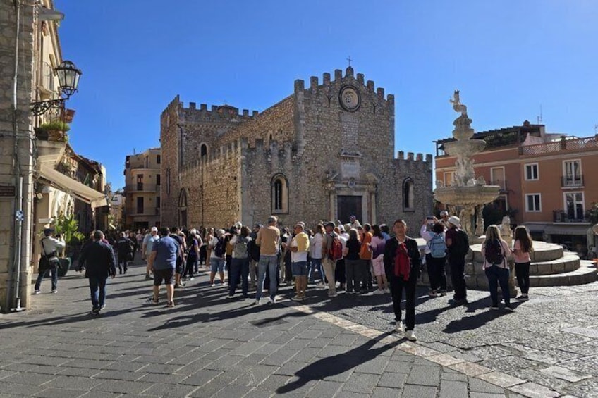 A historic stone building with a tower, a fountain, and a crowd of people in a square. 5
