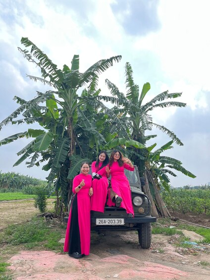 Three individuals in pink dresses seated on a vehicle with a license plate '26A-203.39' in a rural setting with banana trees. 4