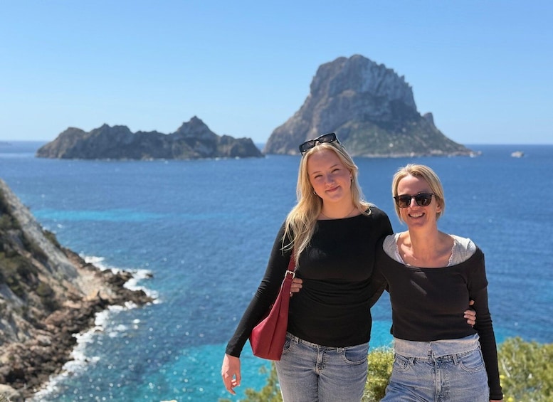Two people standing by a coastal cliff with a clear view of the ocean and a prominent rocky island in the background. 1