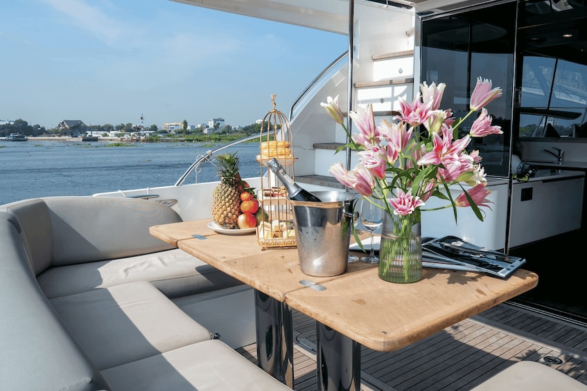 A boat deck with a cushioned seating area, a wooden table with fruits, a metal bucket, and a vase of flowers. 3