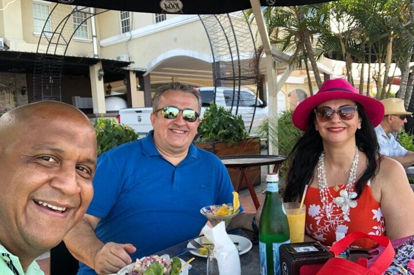 Three people dining outdoors at a restaurant with a tropical setting, including palm trees and a building in the background. 3