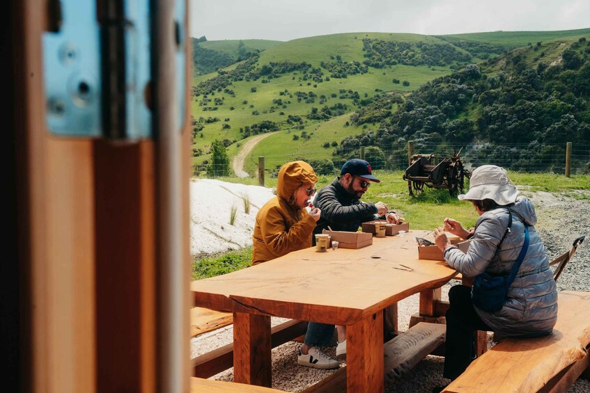 A group of people sitting at a wooden table outdoors with a scenic view of green hills in the background. 4