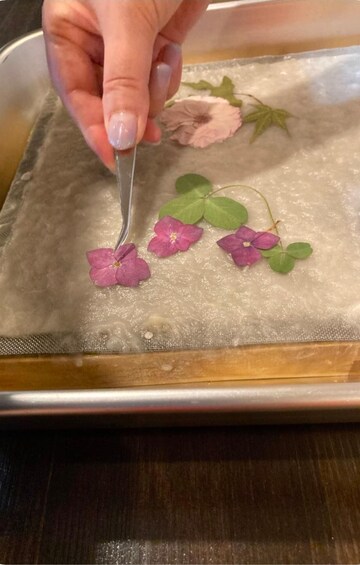 A hand holding a tool over a tray with fabric and purple flowers. 3