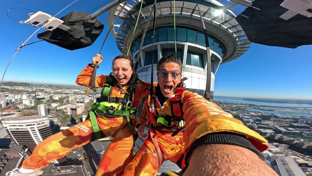 Dos personas hacen bungee jumping desde una estructura alta, con un paisaje urbano y una masa de agua al fondo. 3