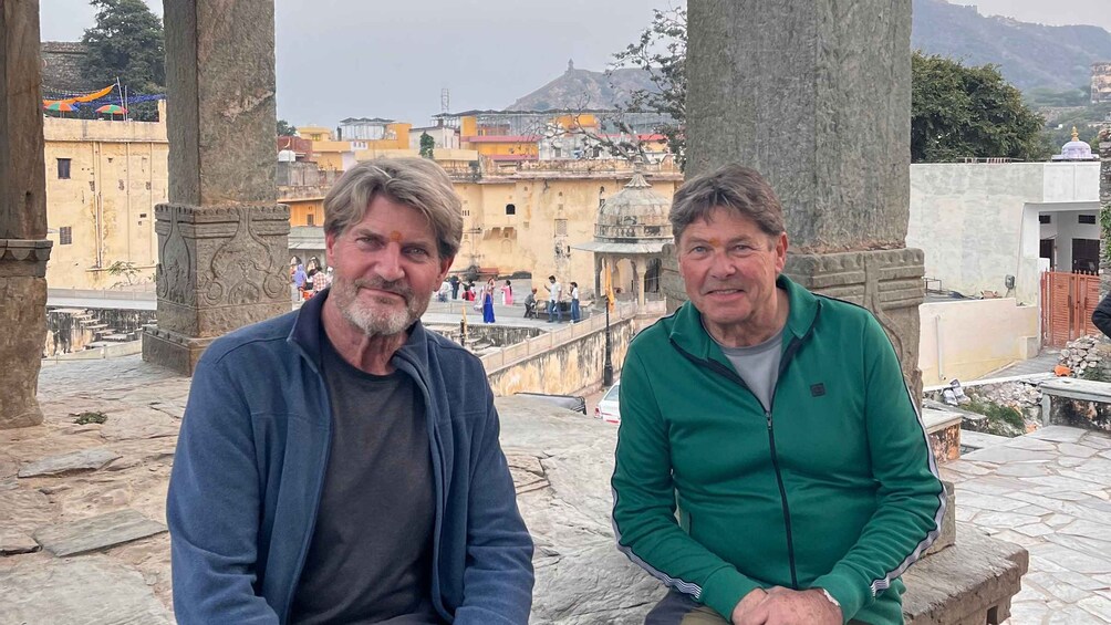 Two men sitting on a stone bench in a historical area with ancient architecture and a view of a cityscape in the background. 2