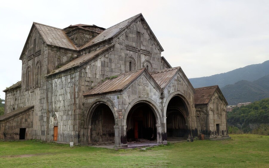 An old stone church with multiple arches and a steep roof, surrounded by a grassy field and mountains in the background. 5