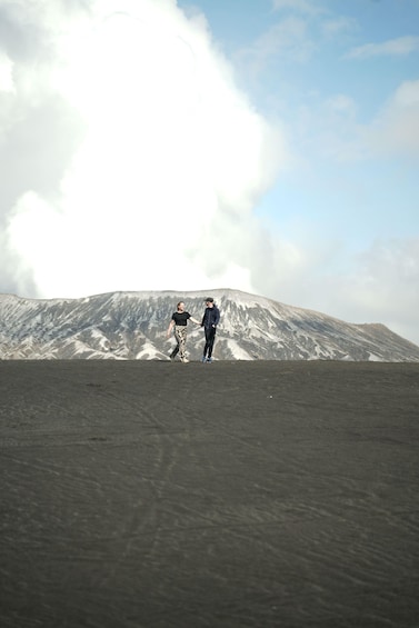 Two people walking on a volcanic landscape with a large cloud in the sky. 2