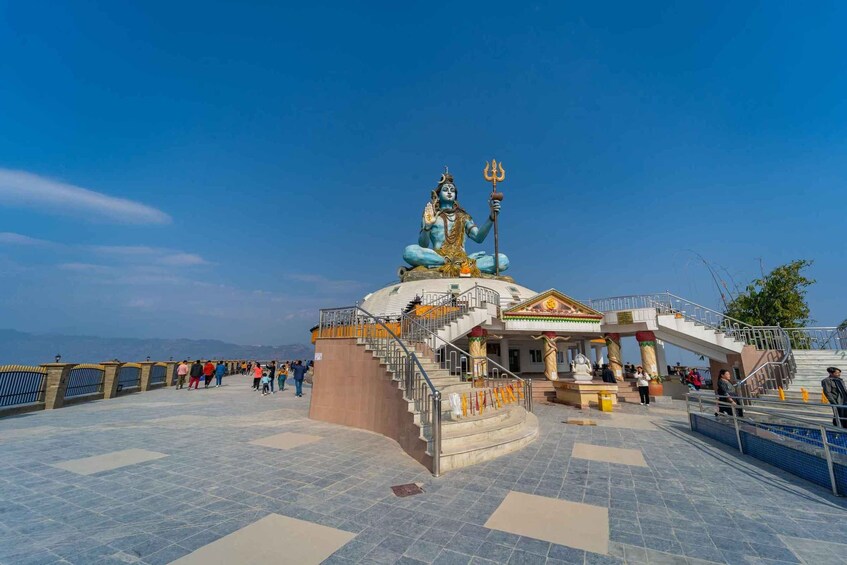 A large statue of a deity with a trident, surrounded by a temple complex with stairs and visitors. 4
