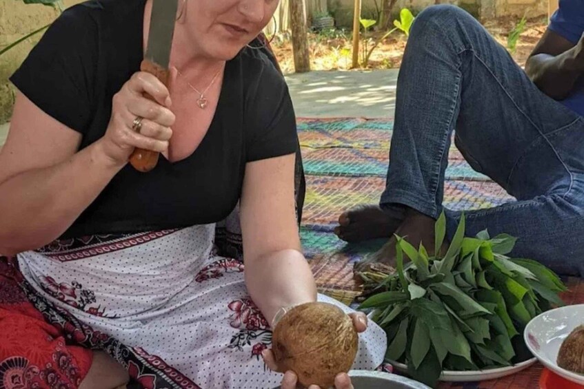 A person holding a coconut and sitting on a mat with a plate of green leaves. 4