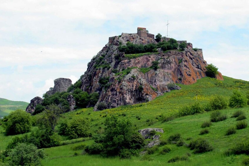 A rocky hill with a small fortress on top, surrounded by green grass and trees. 5