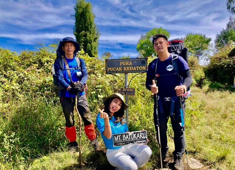 Three people standing in front of a sign that reads "PURA PUCAK KEDATON" and "Mt. Batukaru". 1