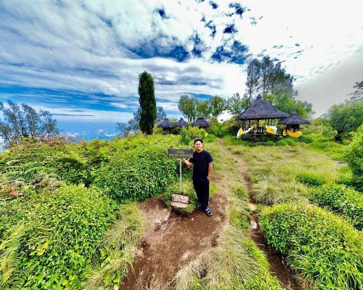 A person standing on a path surrounded by lush greenery, with a thatched-roof structure in the background. 5