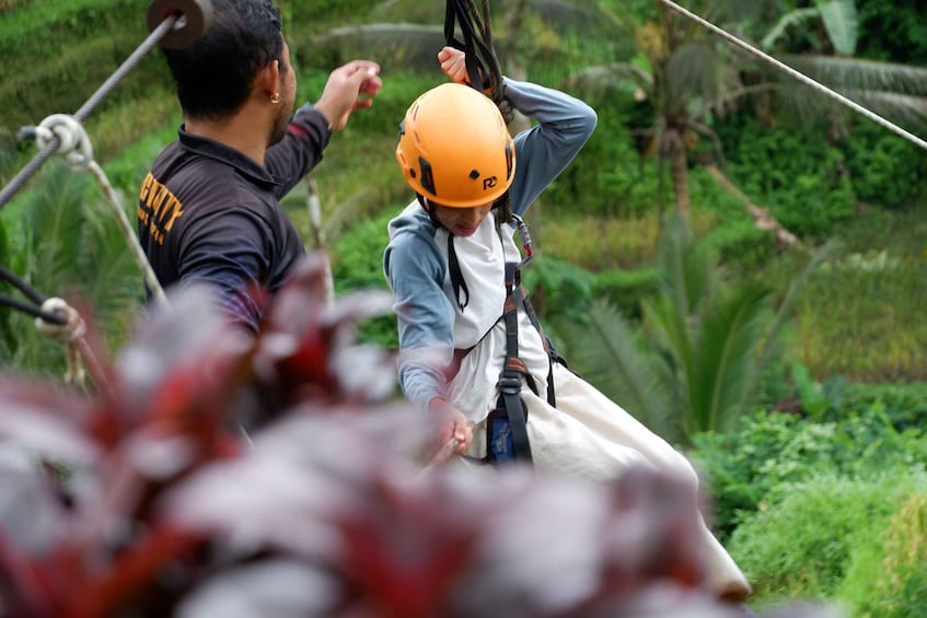 A person in a helmet and harness on a zipline, with a guide assisting. 2