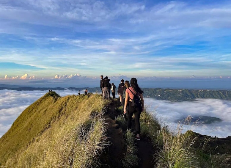 A group of hikers on a mountain trail with a view of clouds below and distant mountains. 1