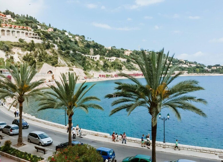 A coastal road with palm trees, a beach, and buildings on a hillside. 2