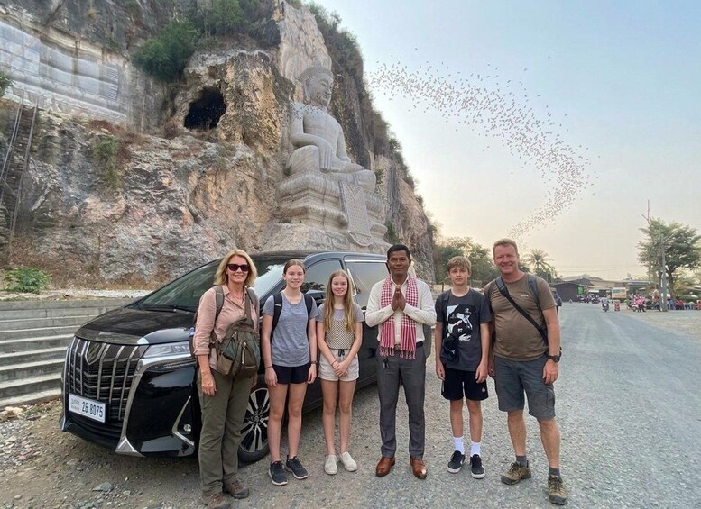 A group of people standing in front of a large rock formation with a statue, a car parked nearby, and a road with a few vehicles in the background. 1