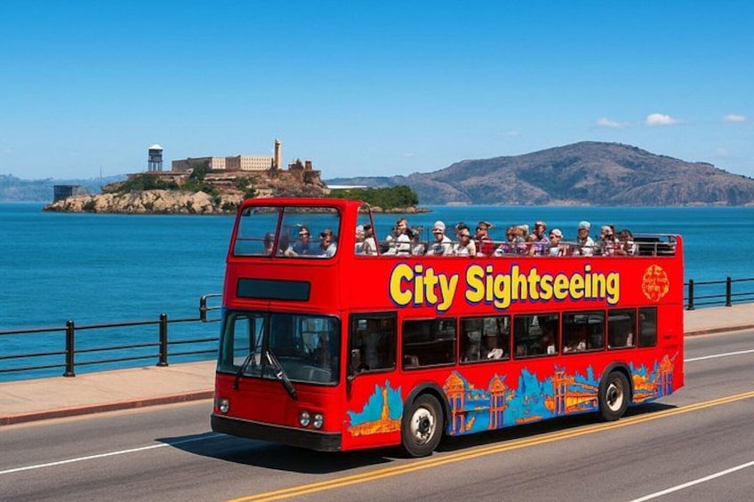 Un autobús rojo de dos pisos con la leyenda 'City Sightseeing' en el lateral, circulando por una carretera con el mar de fondo. 5