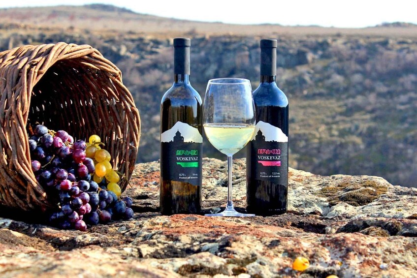 A basket of grapes, two bottles of wine, and a wine glass on a rocky surface with a mountainous background. 5