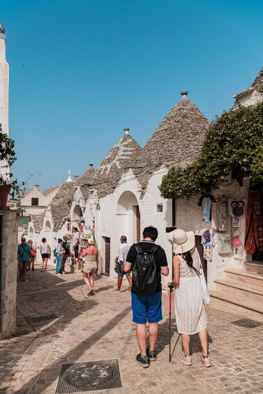A narrow cobblestone street lined with white buildings with conical roofs, people walking, and a man with a walking stick. 2