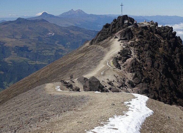A rocky mountain peak with a cross at the summit, a trail leading up, and snow patches. 5