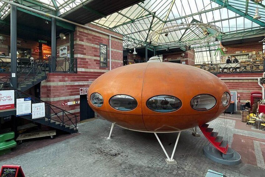 A large orange spherical object with circular windows, displayed indoors with a glass ceiling and brick walls. 5