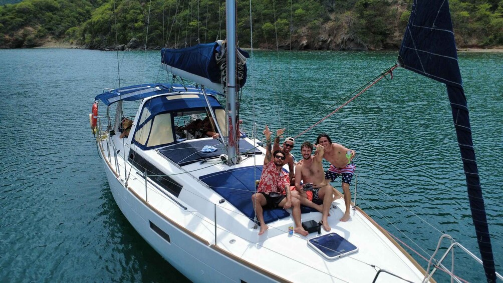 A sailboat with three people on deck, one wearing a red floral shirt and the other two in swimwear, surrounded by calm water and greenery. 4