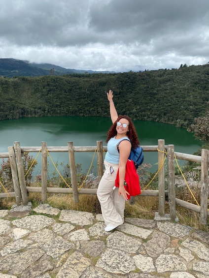 A person standing by a wooden fence overlooking a lake with a forested mountain in the background. 1