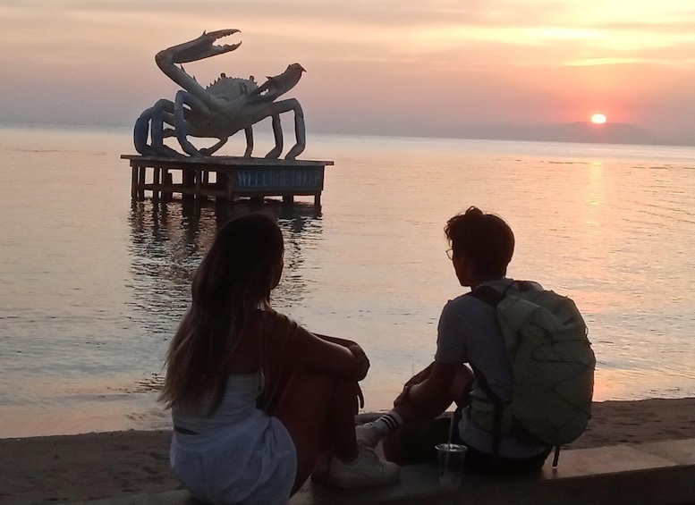 Two people sitting by a beach at sunset, overlooking a sculpture of a crab on a pier. 1