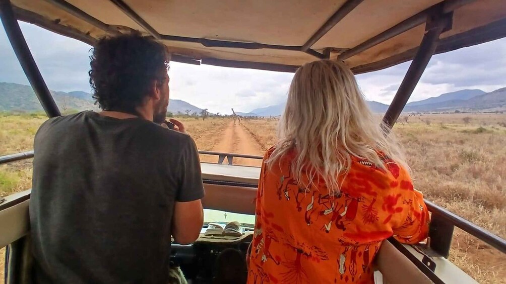 Two people in a safari vehicle driving on a dirt road with mountains in the background. 3