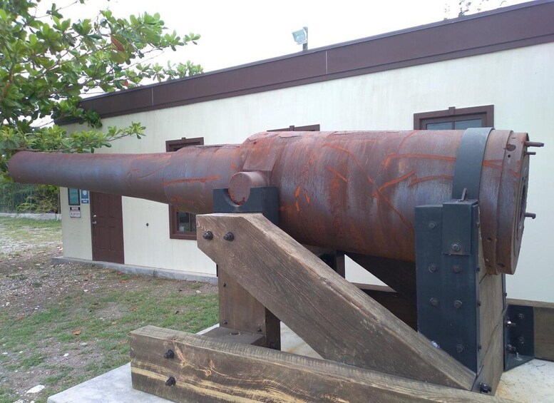 A large, rusted cannon mounted on a wooden platform, with a building in the background. 3