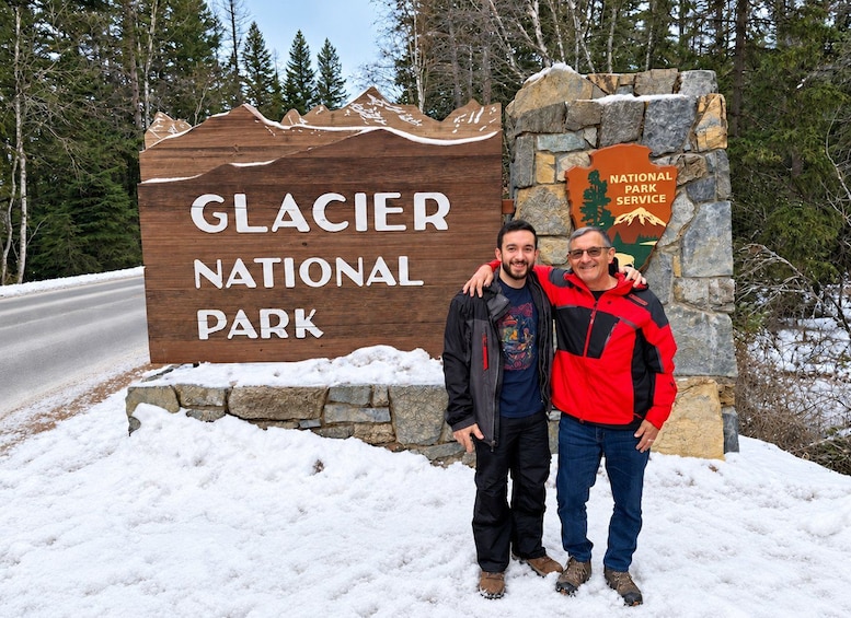 A wooden sign with 'Glacier National Park' written on it, a stone pillar with the National Park Service logo, and two people standing in the snow. 1